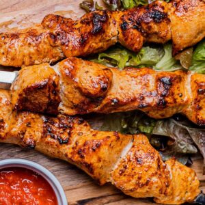 Gegrillte Hähnchenspieße auf einem Holzbrett mit Salatblättern und roter Soße.