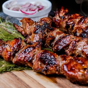 Gegrillte Lammfleischspieße mit Kräutern auf einem Holzbrett und Zwiebelringen im Hintergrund.