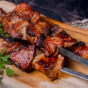 Gegrillte Schweinefleischstücke auf Metallspießen neben Petersilie und kleinen Tomaten.